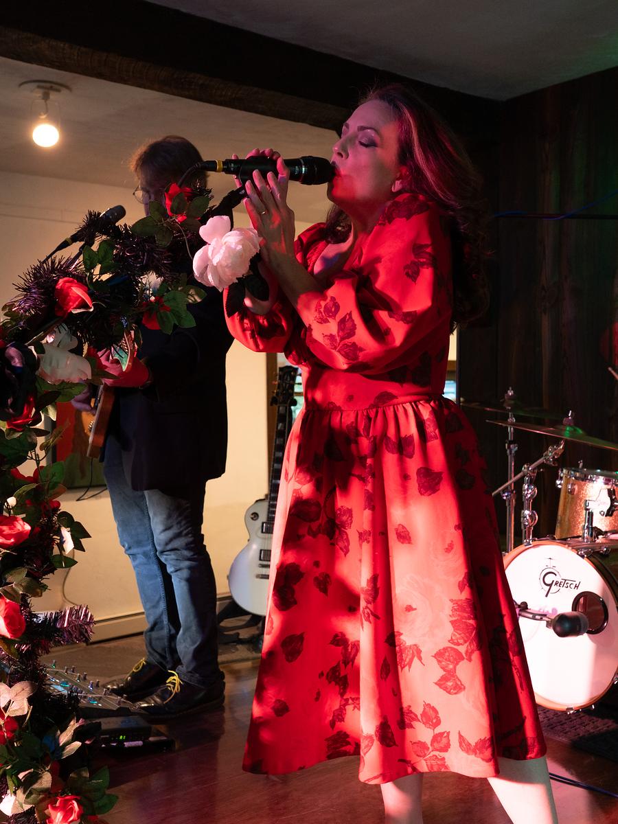 A singer at the microphone under warm red stage light, guitarist and drummer playing behind her
